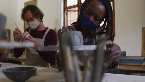 Diverse-male-and-female-potters-wearing-face-mask-and-apron-working-on-pottery-at-pottery-studio