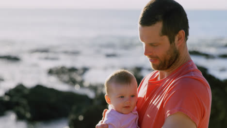 Side-view-of-mid-adult-caucasian-father-holding-baby-at-beach-on-a-sunny-day-4k