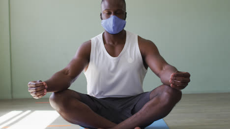 Fit-african-american-man-wearing-face-mask-practicing-yoga-in-yoga-studio