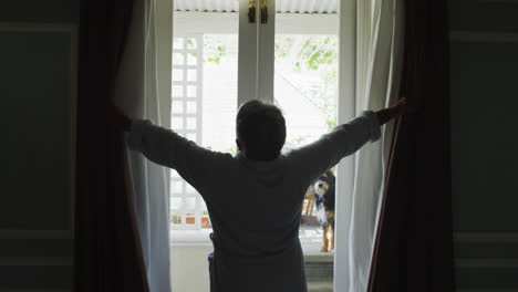 Rear-view-of-senior-mixed-race-woman-at-home-opening-curtains