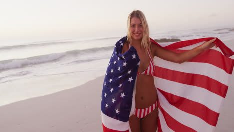 Mujer-Caucásica-Sosteniendo-Y-Ondeando-Una-Bandera-Estadounidense-En-La-Playa.