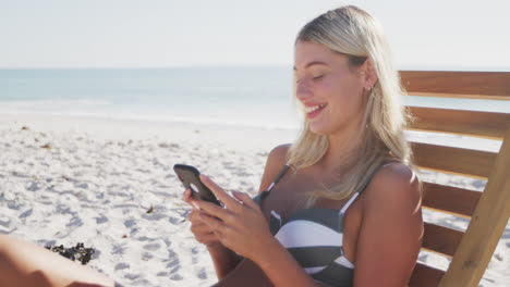 Caucasian-woman-sitting-on-a-sunbed-and-using-her-smartphone-on-the-beach