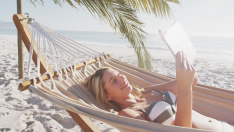 Caucasian-woman-lying-on-a-hammock-and-using-her-tablet-on-the-beach