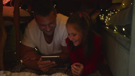 Caucasian-father-and-daughter-lying-and-using-digital-tablet-under-blanket-fort-during-christmas-at-