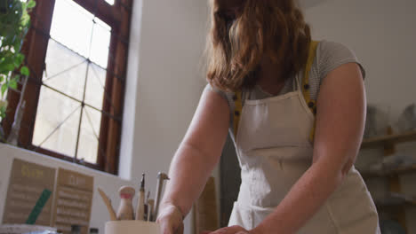 Female-potter-wearing-face-mask-and-apron-kneading-the-clay-at-pottery-studio