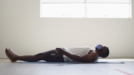 Hombre-Afroamericano-En-Forma-Con-Mascarilla-Practicando-Yoga-Mientras-Está-Acostado-En-Un-Estudio-De-Yoga