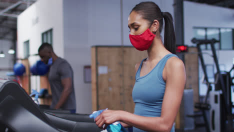 Fit-caucasian-woman-wearing-face-mask-cleaning-treadmill-machine-with-disinfectant-in-the-gym