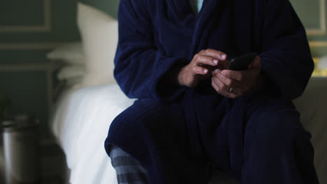 Senior-mixed-race-man-sitting-on-bed-using-smartphone,-midsection