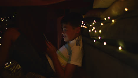 Caucasian-boy-using-smartphone-while-sitting-under-blanket-fort-during-christmas-at-home