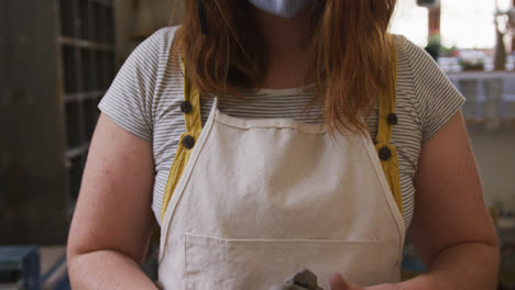 Portrait-of-female-potter-wearing-face-mask-and-apron-working-on-clay-at-pottery-studio