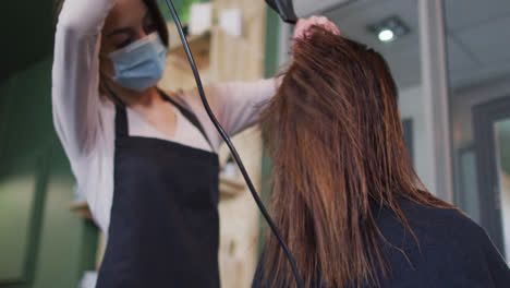 Female-hairdresser-wearing-face-mask-blow-drying-hair-of-female-customer-at-hair-salon