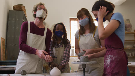 Diverse-potters-wearing-face-masks-having-a-group-video-chat-while-creating-pottery-on-potters-wheel