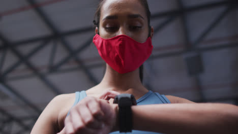 Mixed-race-woman-wearing-face-mask-using-smartwatch-at-gym