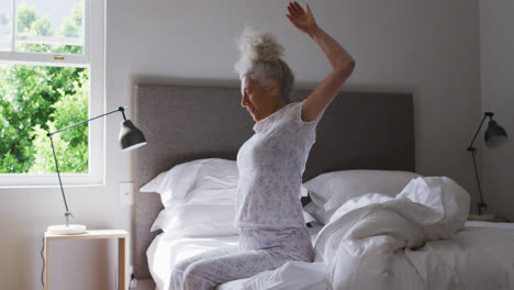 Senior-caucasian-woman-stretching-her-arms-while-sitting-on-the-bed-at-home