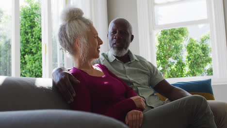 Mixed-race-senior-couple-taking-to-each-other-while-sitting-on-the-couch-at-home