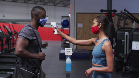 Fit-Kaukasische-Frau-Trägt-Gesichtsmaske-Misst-Temperatur-Von-Fit-Afroamerikanischen-Mann-Im-Fitnessstudio
