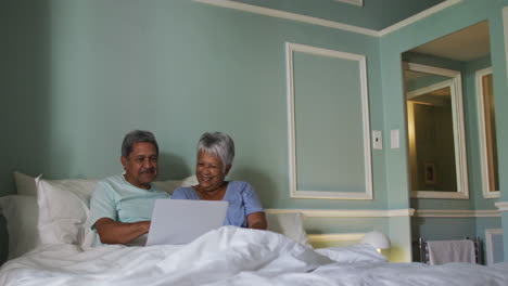 Glückliches-älteres-Mischlingspaar-Sitzt-Im-Bett-Mit-Laptop