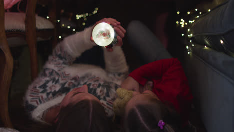 Caucasian-mother-and-daughter-holding-snow-globe-while-lying-under-blanket-fort-during-christmas-at-