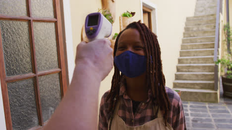 Female-african-american-potter-wearing-face-mask-and-apron-getting-her-temperature-measure-before-en