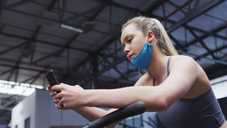 Caucasian-woman-wearing-lowered-face-mask-using-smartphone-at-gym