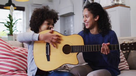 Mixed-race-mother-and-daughter-sitting-on-couch-playing-guitar