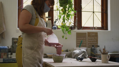 Female-caucasian-potter-wearing-face-mask-pouring-paint-on-pot-at-pottery-studio