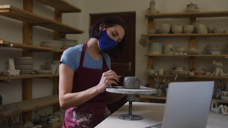 Female-caucasian-potter-wearing-face-mask-using-ribbon-tool-to-create-designs-on-pot-while-having-a-