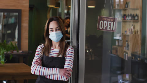 Portrait-of-Female-hairdresser-wearing-face-mask-leaning-on-glass-door-at-hair-salon