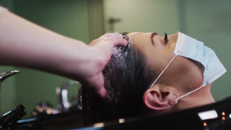 Female-hairdresser-washing-hair-of-female-customer-wearing-face-mask-at-hair-salon