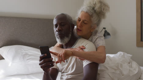 Mixed-race-senior-couple-using-smartphone-while-sitting-on-the-bed-at-home