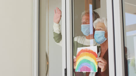 Senior-caucasian-couple-wearing-face-masks-holding-rainbow-painting-against-the-window-at-home