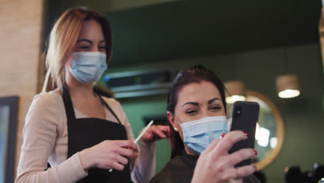 Female-hairdresser-and-female-customer-wearing-face-masks-looking-at-smartphone-at-hair-salon