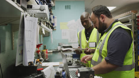 A-Caucasian-and-an-African-American-male-factory-workers-standing,-in-discussion,-using-a-computer-l