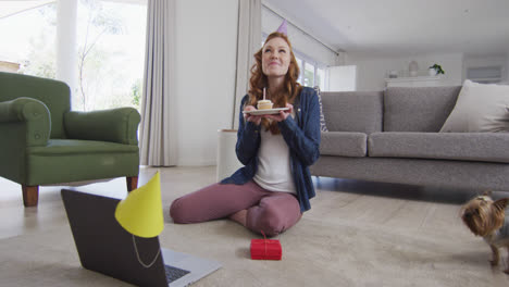 Frau-Mit-Partyhut-Bläst-Kerze-Auf-Den-Kuchen,-Während-Sie-Zu-Hause-Einen-Videochat-Auf-Dem-Laptop-Führt