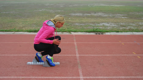 Side-view-of-Caucasian-female-athlete-taking-starting-position-on-running-track-at-sports-venue-4k