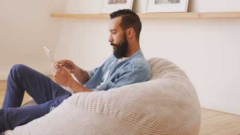 Thoughtful-man-using-digital-tablet-while-relaxing-on-a-bean-bag-indoors