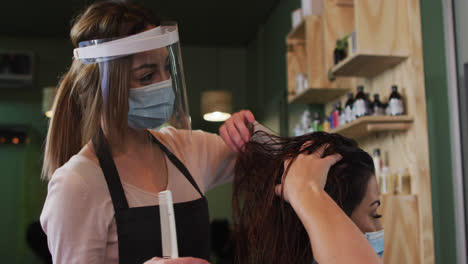 Female-hairdresser-wearing-face-cover-combing-hair-of-female-customer-at-hair-salon