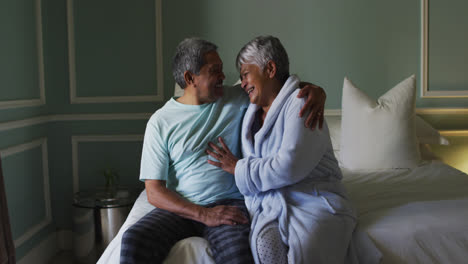 Happy-senior-mixed-race-couple-sitting-on-bed-embracing
