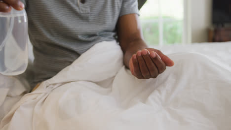 Mittelteil-Eines-Afroamerikanischen-älteren-Mannes,-Der-Seine-Hände-Desinfiziert,-Während-Er-Zu-Hause-Auf-Dem-Bett-Sitzt
