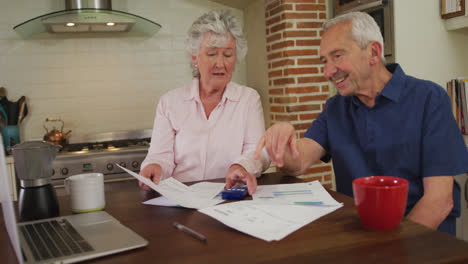 Senior-caucasian-couple-sitting-in-kitchen-checking-paperwork-and-using-laptop-computer-in-slow-moti
