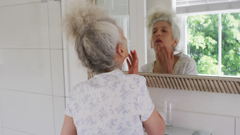Kaukasische-Seniorin-Berührt-Ihr-Gesicht,-Während-Sie-Zu-Hause-In-Den-Spiegel-Schaut