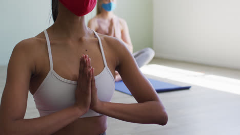 Two-fit-caucasian-women-wearing-face-mask-practicing-yoga-in-yoga-studio