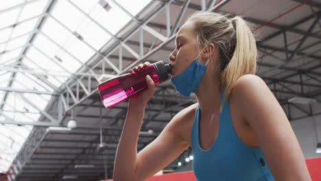Caucasian-woman-wearing-lowered-face-mask-drinking-water-at-gym