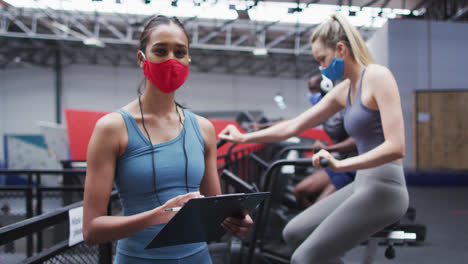 Mixed-race-female-fitness-trainer-wearing-face-mask-at-gym