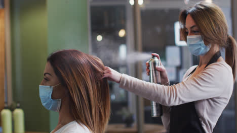 Female-hairdresser-wearing-face-mask-spraying-hairspray-on-hair-of-female-customer-at-hair-salon