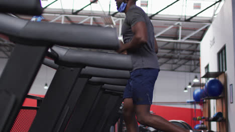 Fit-african-american-man-with-face-mask-around-his-neck-running-on-treadmill-in-the-gym