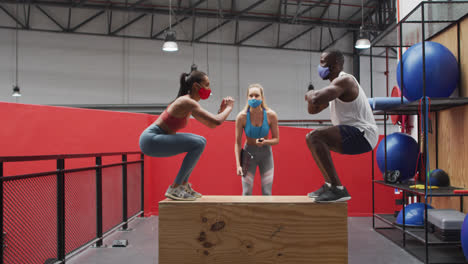 Diverse-man-and-woman-jumping-on-box-wearing-face-masks-at-gym,-caucasian-woman-timing