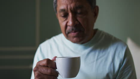 Senior-mixed-race-man-sitting-in-bedroom-drinking-coffee