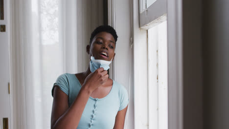 African-american-woman-removing-face-mask-and-looking-out-of-window-at-home