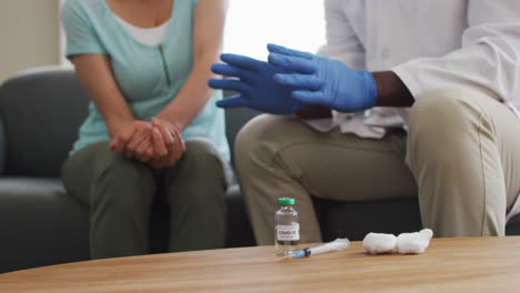 Mid-section-of-male-african-american-doctor-talking-to-female-patient-before-injecting-covid-19-vacc